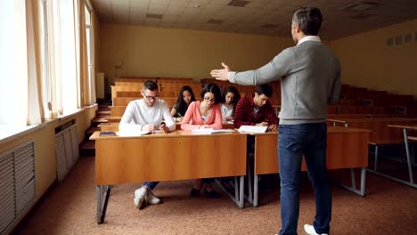 Fleißige-Schüler-sind-Schreibtest-an-Schreibtischen-im-Hörsaal-sitzen,-während-junger-Mann-in-Gläsern-im-Gespräch-mit-Lehrer-Fragen-stellt.-Bildung,-wissen-und-Menschen-Konzept.