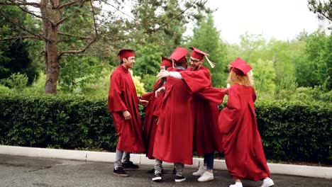 Slow-motion-of-young-men-and-women-grads-shaking-hands,-doing-high-five,-dancing-and-hugging-outdoors-on-dimmer-day-with-beautiful-nature-in-background.