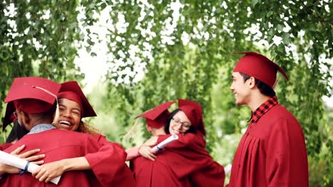 Happy-graduating-students-multi-ethnic-group-is-hugging-and-doing-high-five-after-graduation-ceremony,-girls-and-guys-in-mortar-boards-and-gowns-are-laughing-and-having-fun.