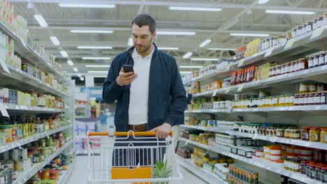 Im-Supermarkt:-gut-aussehend-Mann-nutzt-Smartphone,-lächelt-beim-stehen-im-Abschnitt-aus-der-Dose-waren.-Warenkorb-mit-gesunden-Lebensmitteln-im-Inneren-hat.-Andere-Kunden,-die-im-Hintergrund-laufen.