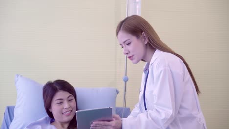 Beautiful-smart-Asian-doctor-and-patient-discussing-and-explaining-something-with-tablet-in-doctor-hands-while-staying-on-Patient's-bed-at-hospital.-Medicine-and-health-care-concept.