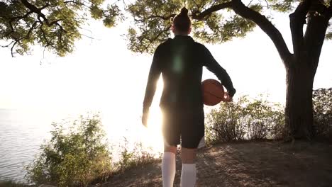 A-basketball-player-girl-comes-with-a-ball-in-her-hand,-coming-up-to-the-slope-with-trees-around.-Looks-at-the-sun-shining-over-the-sea.-Backside-view