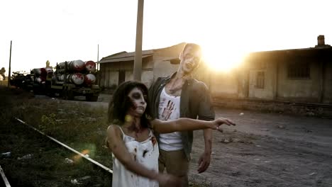 Halloween,-filming,-creepy-concept.-Creepy-zombie-man-and-woman-in-bloody-clothes-walking-by-railway-lines-outdoors-by-industrial-abandoned-place.-Sun-shines-on-the-background