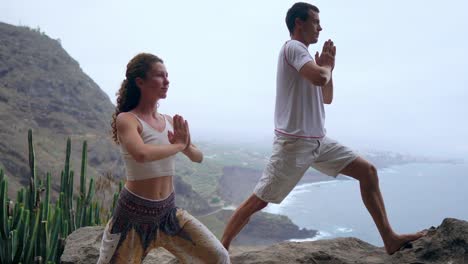 Un-hombre-y-una-mujer-de-pie-en-el-borde-de-un-acantilado-con-vistas-al-océano-levantan-las-manos-y-aspirar-el-aire-del-mar-durante-el-yoga.