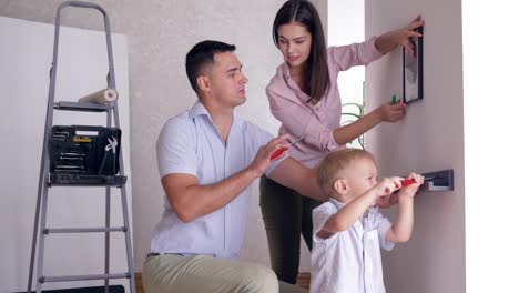 freundliche-Familie-Reparaturen-ausführen-und-kleinen-Sohn-hilft-Eltern-Regal-in-neue-Wohnung-an-Wand-befestigen