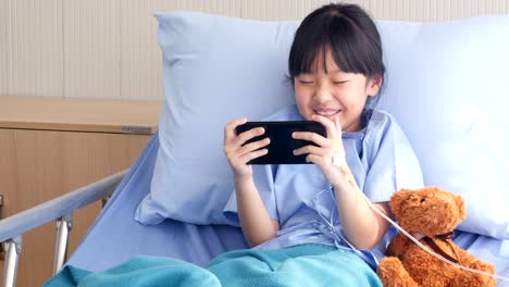 Kleine-Mädchen-liegt-auf-einem-Bett-im-Krankenhaus,-sie-Filme/Zeichentrickfilme/lustige-Videos-auf-Smartphone.-Menschen-mit-Technik,-Gesundheits--und-medizinische-Konzept.