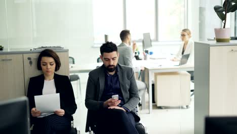 Stressed-candidates-are-waiting-for-job-interview-in-office-reading-cv-and-using-smartphone-while-female-manager-is-interviewing-a-man-in-room.-Company-and-staff-concept.