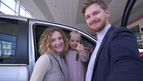 Familienselfi-FotoFoto-im-Autohaus,-lustiges-Paar-mit-niedlichen-Kindermädchen-mit-Schlüsseln-auf-Smartphone-in-der-Nähe-von-neu-gekauftem-Automobil-im-Autoverfieberzentrum-fotografiert