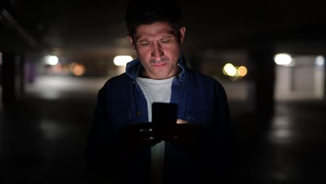 Verdächtig-aussehender-hispanischer-Mann-denkt,-während-er-Telefon-auf-dunklem-Parkplatz-benutzt