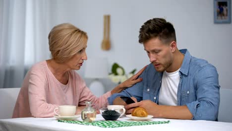 Worried-mother-talking-to-infantile-adult-son-playing-video-games-on-smartphone