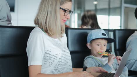 Mamá-joven-trabajando-en-la-computadora-portátil-en-el-aeropuerto-con-el-niño-disfrutando-de-piruleta.