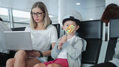 Junge-Mutter-arbeitet-auf-Laptop-am-Flughafen-mit-Kind-genießen-Lutscher.