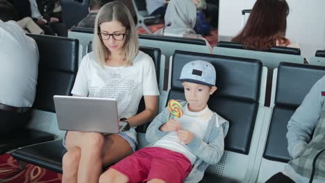 Mamá-joven-trabajando-en-la-computadora-portátil-en-el-aeropuerto-con-el-niño-disfrutando-de-piruleta.