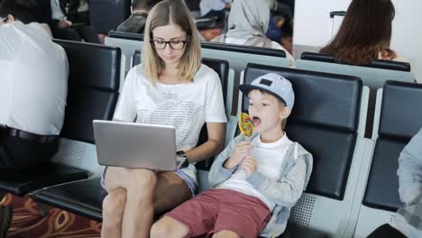 Junge-Mutter-arbeitet-auf-Laptop-am-Flughafen-mit-Kind-genießen-Lutscher.