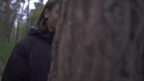 Bonita-chica-con-abrigo-caliente-mirando-desde-detrás-de-un-árbol-sonriendo.-La-cámara-se-mueve-a-otro-lado-del-maletero