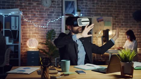 Man-using-virtual-reality-glasses-moving-arms-sitting-at-desk-in-dark-office