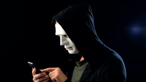 Anonymous-enmascarado-estafador-en-la-capucha-exige-un-rescate-para-el-chantaje-utilizando-un-teléfono-inteligente.-Masked-Criminal-intimida-a-la-víctima-con-la-ayuda-de-amenazas-a-través-de-SMS-utilizando-el-teléfono-móvil.