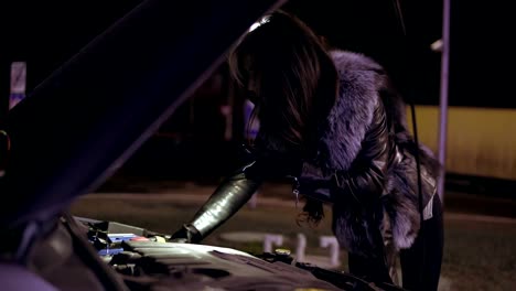 Young-brunette-woman-with-long-hair-is-looking-under-the-hood-using-her-smartphone-as-a-flashlight