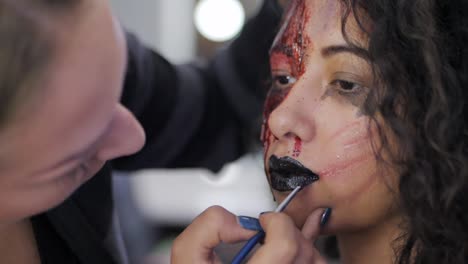 Make-up-artist-make-the-girl-halloween-make-upin-studio.Halloween-face-art.-Woman-applies-on-black-lipstick-with-brush-on-lips-of-latin-girl.-War-paint-with-blood,-scars-and-wounds.Slow-motion