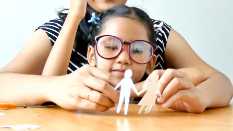 Cerrar-tiro-niña-asiática-en-estudiante-de-kindergarten-tailandés-uniforme-y-a-su-madre-jugar-corte-de-papel-blanco-que-forma-familia-padre-madre-hijo-e-hija-en-la-mesa-de-madera-seleccione-profundidad-de-foco-de-campo