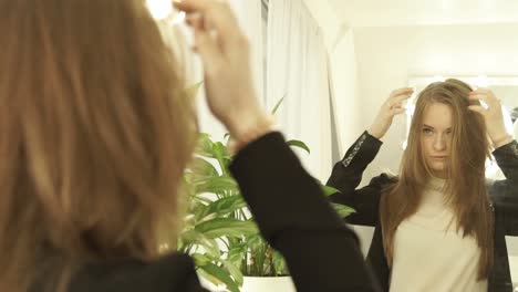 Hermosa-mujer-tocando-pelo-y-mirando-en-el-espejo-en-el-salón-de-peluquería.-Joven-rubia-mirando-el-peinado-en-el-salón-de-belleza-de-cerca