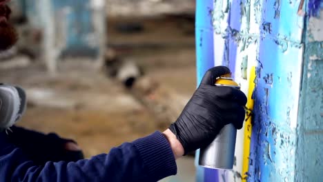 Close-up-shot-of-male-hand-in-protective-glove-holding-aerosol-paint-and-painting-graffiti-on-pillar-inside-abandoned-building.-Modern-art,-abstract-images-and-people-concept.