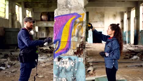 Slow-motion-of-two-serious-graffiti-artists-decorating-empty-industrial-building-with-beautiful-images-using-bright-aerosol-paint.-Creative-teamwork,-people-and-art-concept.