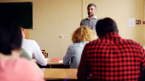 High-school-teacher-handsome-man-is-talking-to-students-who-are-sitting-at-tables-in-classroom-and-smiling.-Education,-teaching-and-learning,-people-concept.