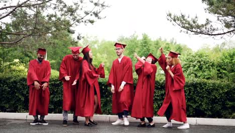 Slow-motion-of-multi-ethnic-group-of-students-in-gowns-and-mortar-boards-dancing-and-laughing-outdoors-on-campus-on-graduation-day-celebrating-event.