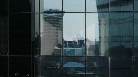 Aerial-Footage-From-Outside-of-the-Skyscraper:-Businessman-and-Businesswoman-Talking-Business-while-Sitting-at-the-Desk-in-the-Office-Window.-Flying-Shot-of-the-Financial-District-and-Businesspeople-Working-in-the-Big-City.