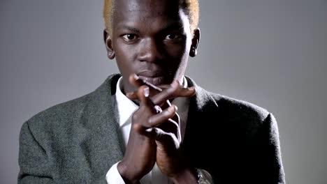 Young-afro-american-blond-man-in-suit-holding-hands-and-praying,-looking-in-camera,-isolated-on-grey-background