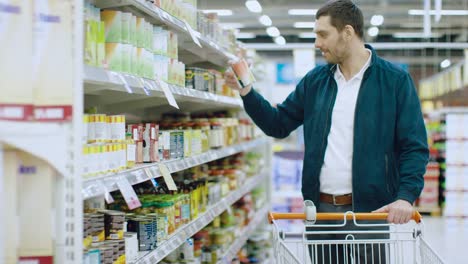 At-the-Supermarket:-Handsome-Man-Browses-Through-Shelf-with-Canned-Goods,-Chooses-Tin-Can-and-Places-it-into-His-Shopping-Cart.