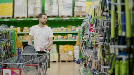 Alone-young-man-is-walking-in-sales-hall-in-shop,-rolling-trolley-in-front