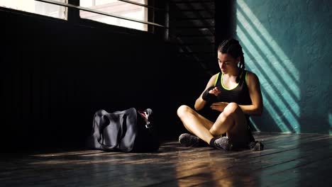 Panning-young-beautiful-woman-sitting-on-floor-and-wrapping-hands-with-black-boxing-wraps-in-club.