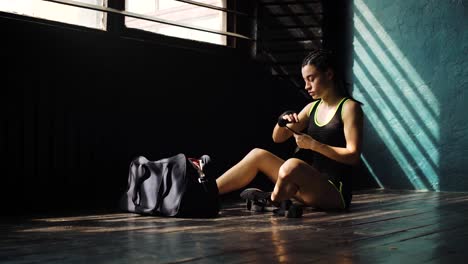 Panning-young-beautiful-woman-sitting-on-floor-and-wrapping-hands-with-black-boxing-wraps-in-club.