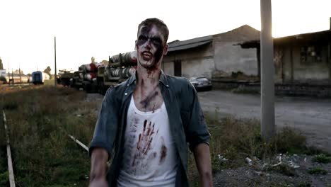 Creepy-zombie-man-in-bloody-clothes-walking-by-railway-lines-outdoors-with-an-industrial-abandoned-place-on-the-background.-Halloween,-filming,-creepy-concept