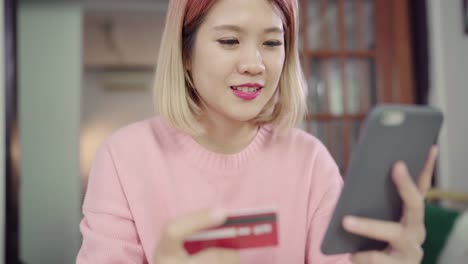 Beautiful-Asian-woman-using-smartphone-buying-online-shopping-by-credit-card-while-wear-sweater-sitting-on-desk-in-living-room-at-home.-Lifestyle-woman-at-home-concept.