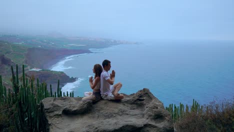 Mann-und-Frau-sitzen-auf-einem-Berg-auf-einem-Felsen-zurück-zu-meditieren-und-Yoga-auf-dem-Hintergrund-des-Ozeans-zu-tun.