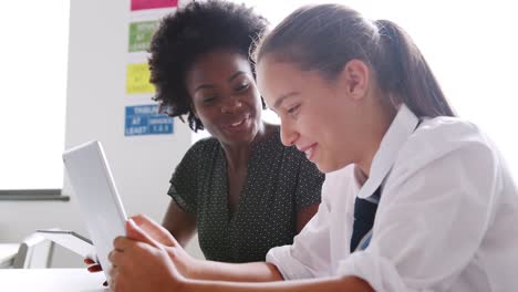 Tutor-de-secundaria-mujer-con-tableta-Digital-dando-a-chica-estudiante-con-uniforme-enseñanza-uno-a-uno-en-el-escritorio