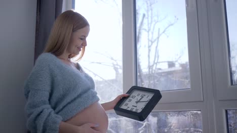 Baby-erwartet,-hält-junge-schwangere-Frau-mit-großen-Bauch-Wanduhr-in-der-hand-gegen-Fenster-im-Sonnenlicht-an-Wintertag