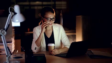 Attractive-female-entrepreneur-is-talking-on-mobile-phone-and-using-laptop-working-in-office-late-at-night.-Modern-technology,-hard-working-youth-and-communication-concept.