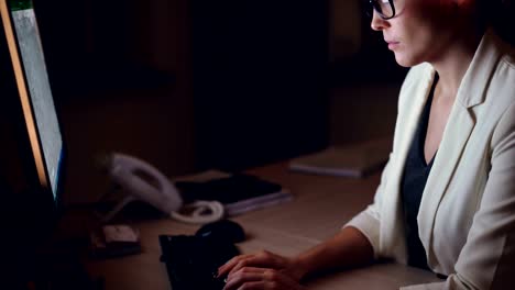 Tilt-up-shot-of-serious-young-woman-working-on-computer-typing-and-looking-at-screen-sitting-at-worktable-alone-in-dark-office-late-at-night.-Technology-and-job-concept.