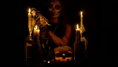 Bruja-de-Halloween-con-maquillaje-de-cráneo-hace-vudú-sostiene-cuchillo-y-wispering-hechizo-mágico-cadenas-de-calabaza-y-velas