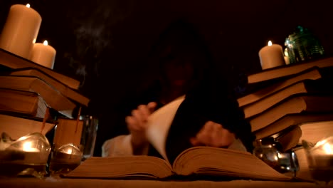 Medium-close-up-girl-magician-in-a-hood-in-a-dark-room-by-candlelight-and-looking-for-a-spell-turning-over-a-book.-Low-key.