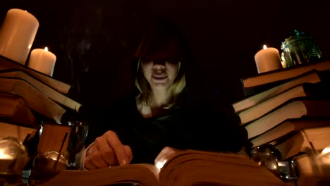 Medium-close-up-girl-magician-in-a-hood-in-a-dark-room-by-candlelight-and-looking-for-a-spell-turning-over-a-book.-Low-key.