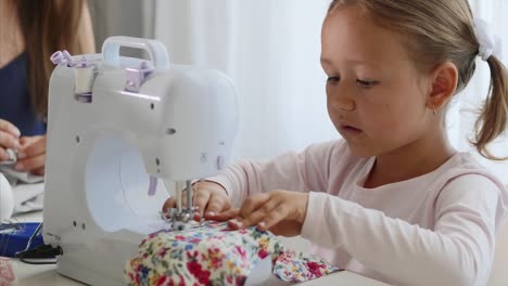 A-little-girl-is-sewing-fabric-and-talking.-Mom-is-at-the-background.-Close-up.