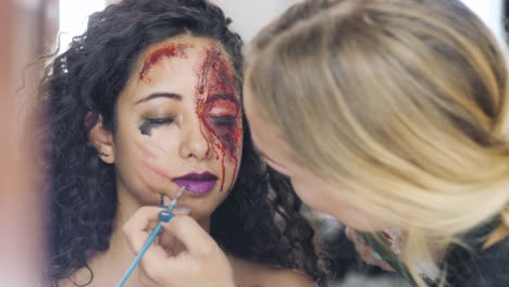 Make-up-artist-make-the-girl-halloween-make-upin-studio.Halloween-face-art.Woman-applies-on-professional-greasepaint-on-the-face-of-spanish-girl.War-paint-with-blood,-scars-and-wounds.-4k