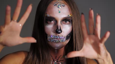 Chica-joven-con-arte-creativo-de-cara-halloween-asustar-en-el-estudio.-Aterrador-retrato-de-glamour-cráneo-con-pedrería-y-lentejuelas.-Maquillaje-profesional-para-la-celebración.-Cámara-lenta