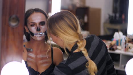 Artista-de-maquillaje-pinturas-de-maquillaje-para-Halloween-en-estudio.-Mujer-dibujando-una-calavera-glamorosa-con-pedrería-y-lentejuelas-en-una-hermosa-joven-con-el-pelo-largo.-Reflexión-en-el-movimiento-de-Mirror.Slow