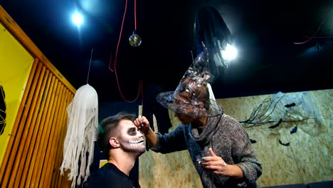 Halloween-Party,-Make-up-Artist-zieht-eine-schreckliche-Make-up-auf-dem-Gesicht-eines-Mannes-für-eine-Halloween-Party.-im-Hintergrund-sieht-man-die-Landschaft-im-Stil-von-Halloween